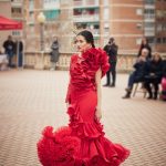 Vestido de flamenca rojo con encaje asimétrico con conjunto de volantes a un lado y manga larga de encaje al otro. En la parte inferior un volante con subida central y un volante con mucho volumen en forma canastera, además lleva una gran enagua de rizo para crear más volumen