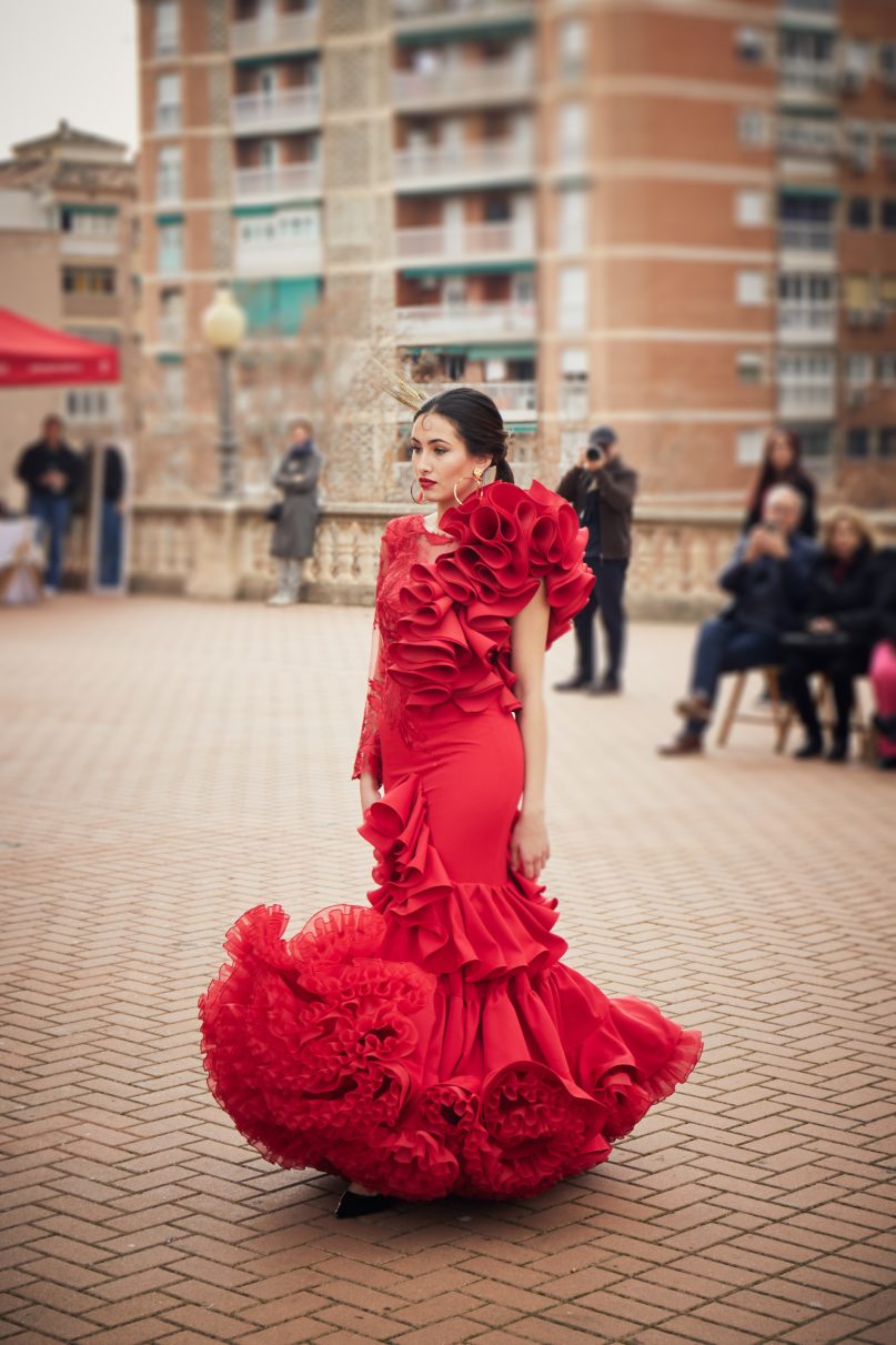 Vestido de flamenca rojo con encaje asimétrico con conjunto de volantes a un lado y manga larga de encaje al otro. En la parte inferior un volante con subida central y un volante con mucho volumen en forma canastera, además lleva una gran enagua de rizo para crear más volumen