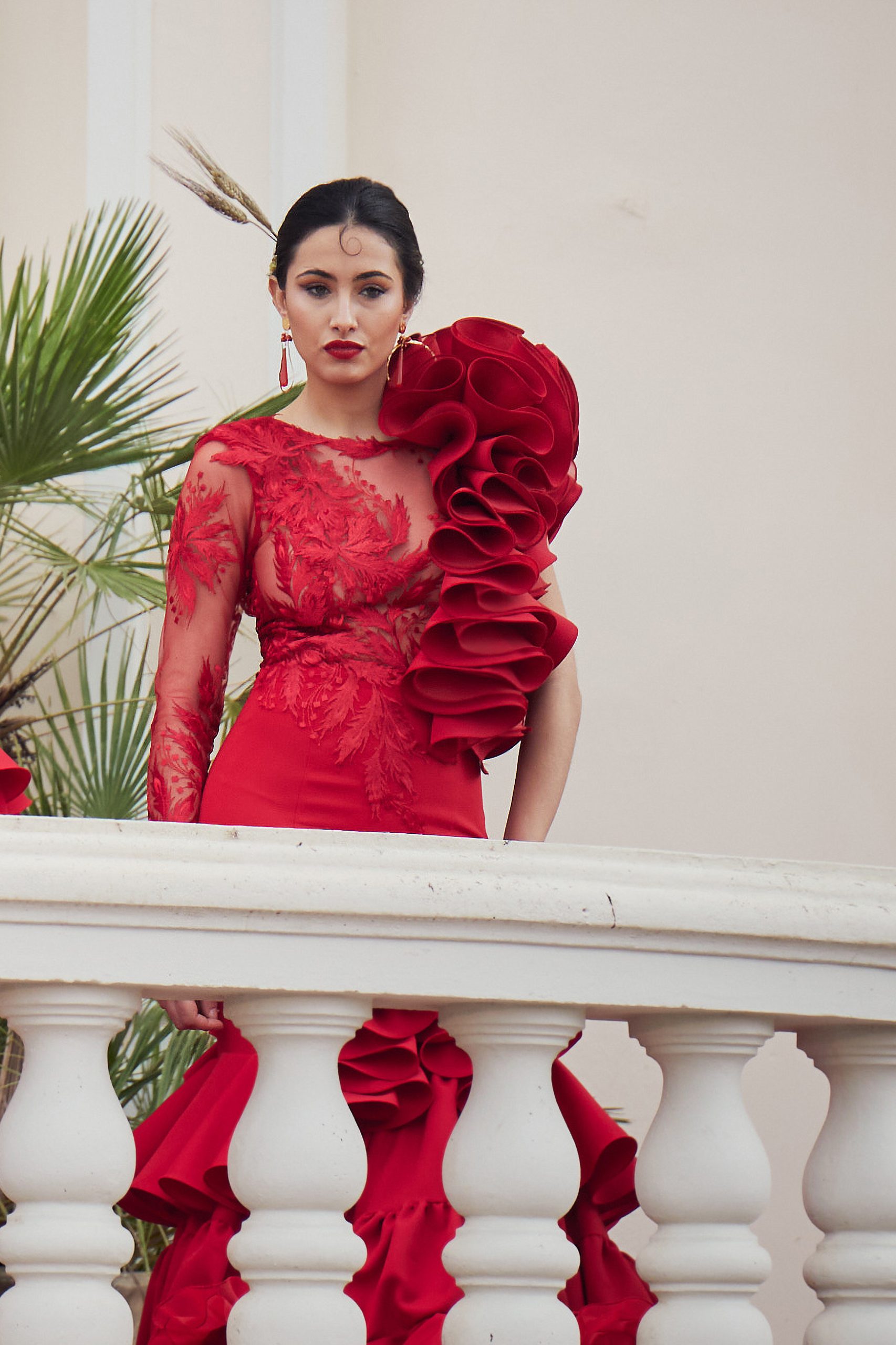 Vestido de flamenca rojo con encaje asimétrico con conjunto de volantes a un lado y manga larga de encaje al otro. En la parte inferior un volante con subida central y un volante con mucho volumen en forma canastera, además lleva una gran enagua de rizo para crear más volumen