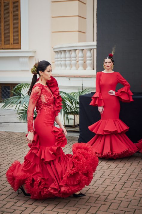 Vestido de flamenca rojo con encaje asimétrico con conjunto de volantes a un lado y manga larga de encaje al otro. En la parte inferior un volante con subida central y un volante con mucho volumen en forma canastera, además lleva una gran enagua de rizo para crear más volumen