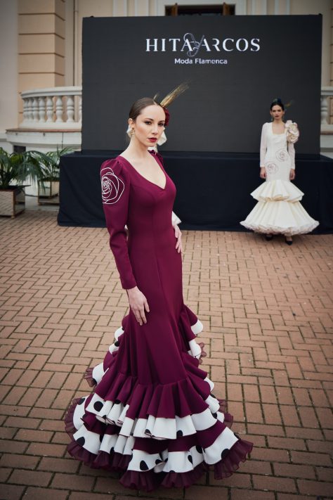 Vestido flamenca de nesgas en color cardenal con bordado de rosa en crudo en ambas mangas en la copa. Es un vestido con dos dobles volantes en la parte inferior uno en color cardenal y el interior en color crudo con lunar negro galleta. El volante superior tiene una subida muy favorecedora en uno de los costados.