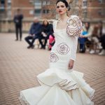 Vestido flamenca entallado en color crudo con bordado de rosa en granate, escote cuadrado y mangas de gasa. Es un vestido con dos volantes en la parte inferior y con un juego de volantes a un hombro creando volumen en la partes superior de la manga.
