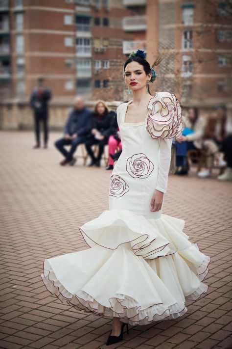 Vestido flamenca entallado en color crudo con bordado de rosa en granate, escote cuadrado y mangas de gasa. Es un vestido con dos volantes en la parte inferior y con un juego de volantes a un hombro creando volumen en la partes superior de la manga.