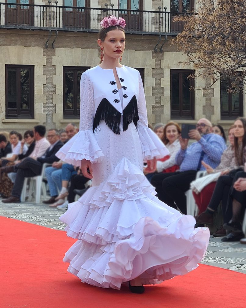 Vestido flamenca blanco tejido perforado con tres volantes, el primero con subida en el delantero todos dobles con rizo en popelín, escote en pico delantero y a la caja trasero. Mangas largas con volante perforado doble con rizo de popelín.