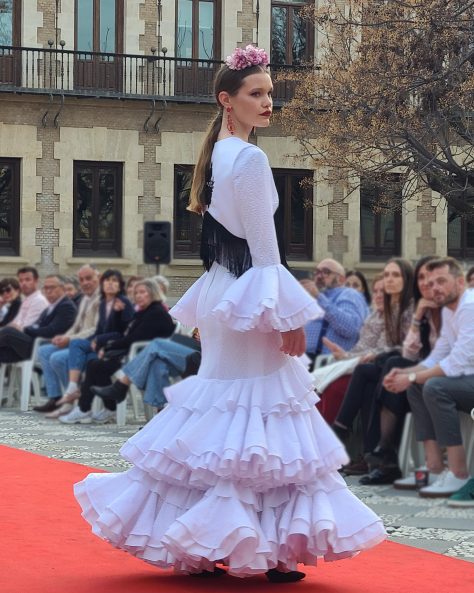 Vestido flamenca blanco tejido perforado con tres volantes, el primero con subida en el delantero todos dobles con rizo en popelín, escote en pico delantero y a la caja trasero. Mangas largas con volante perforado doble con rizo de popelín.