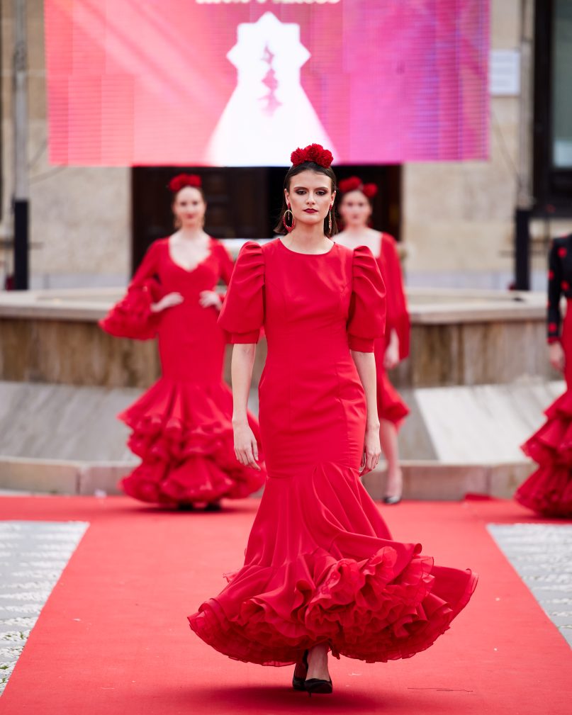 Vestido de flamenca rojo, mangas abullonadas, escote cerrado delantero y abierto en forma triangulo trasero. Volantes grande con mucho volumen y enaguas de rizo en rojo. Detalle encaje en espalda