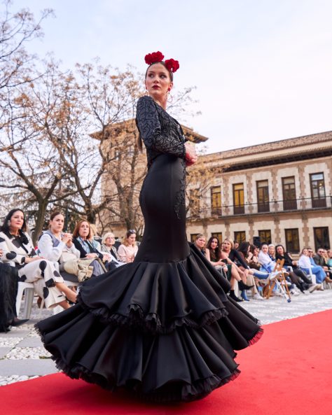 Vestido flamenca en tejido negro satinado con escote redondo por delante y por la espalda profundo, sin mangas con encaje como tirante y adorno en el cuerpo, con dos volantes grandes