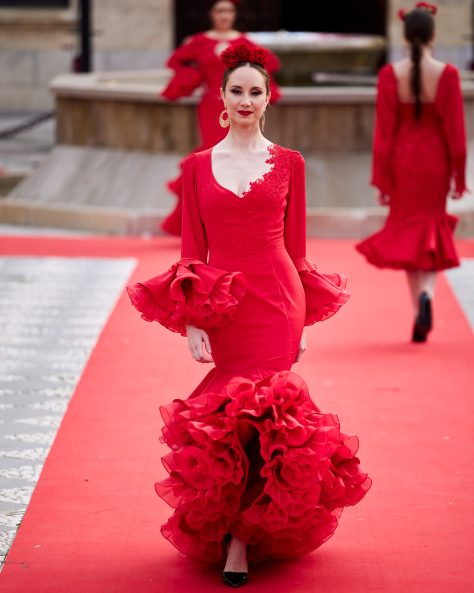 Vestido flamenca rojo con dos volantes, uno asimétrico, mangas amplias con volante de mucho volumen, escote en v con adornos de pasamanería. Volante grande y con rizo en el bajo