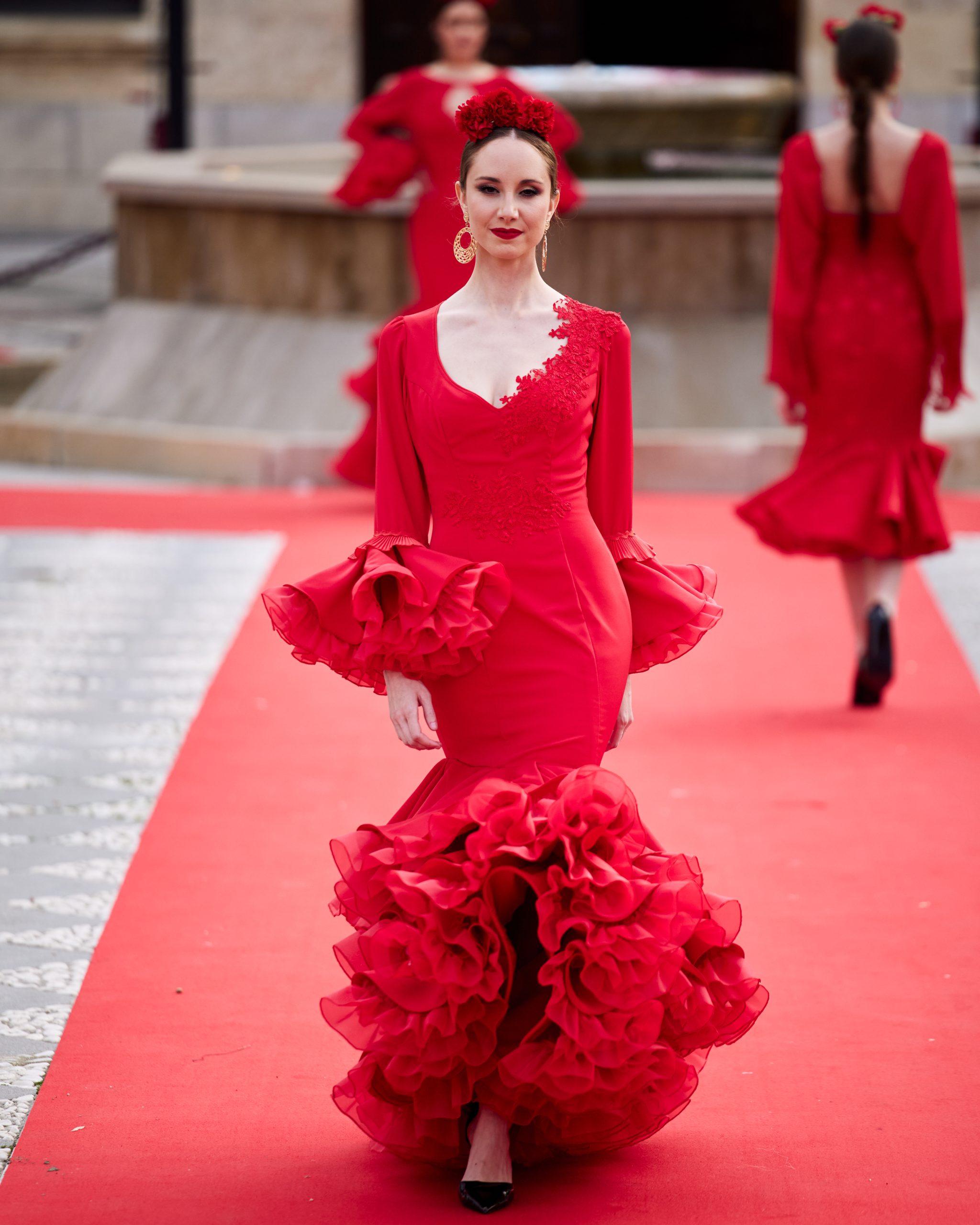 Vestido flamenca rojo con dos volantes, uno asimétrico, mangas amplias con volante de mucho volumen, escote en v con adornos de pasamanería. Volante grande y con rizo en el bajo