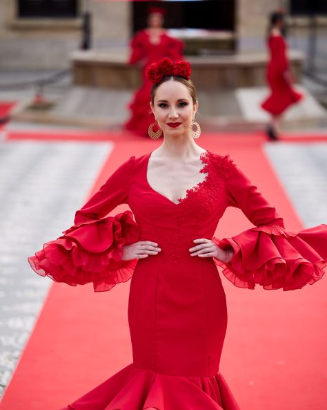 Vestido flamenca rojo con dos volantes, uno asimétrico, mangas amplias con volante de mucho volumen, escote en v con adornos de pasamanería. Volante grande y con rizo en el bajo