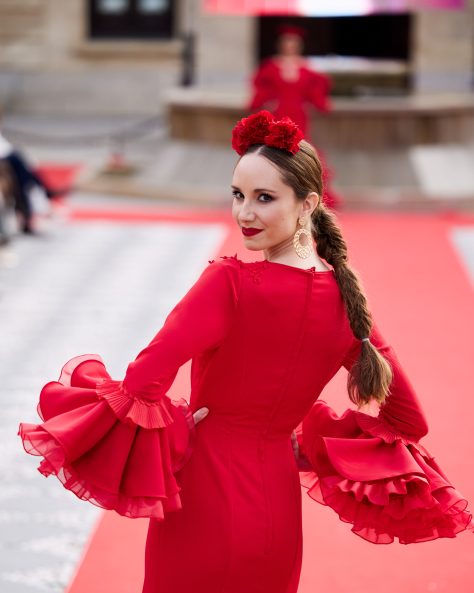 Vestido flamenca rojo con dos volantes, uno asimétrico, mangas amplias con volante de mucho volumen, escote en v con adornos de pasamanería. Volante grande y con rizo en el bajo